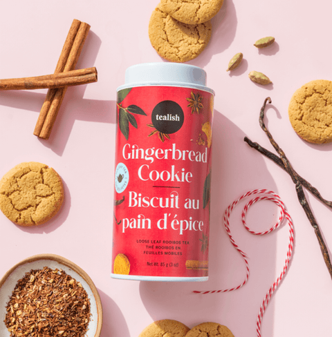 Gingerbread Cookie rooibos tea blend canister surrounded by cookies, spices, and loose tea on a pink background.