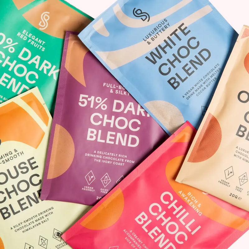 A colorful assortment of Blendsmiths drinking chocolate sachets, featuring flavors like 51% Dark Choc Blend, White Choc Blend, Chilli Choc Blend, and more. Each sachet has a modern design with unique color schemes and highlights key attributes such as veg