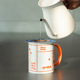 A hand holds a white pour-over kettle above a white ceramic mug with an orange handle and orange desert-themed illustrations, including camels and the text 'just like this'.
