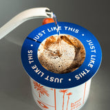 A close-up of a pour-over coffee setup with hot water being poured from a white kettle into a paper filter basket. The basket has a blue rim with white text reading 'JUST LIKE THIS' and a white cup below with orange palm tree illustrations.