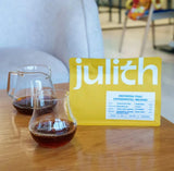 Julith Indonesia Prau Experimental Washed specialty coffee bag with two glasses of brewed coffee on a wooden table