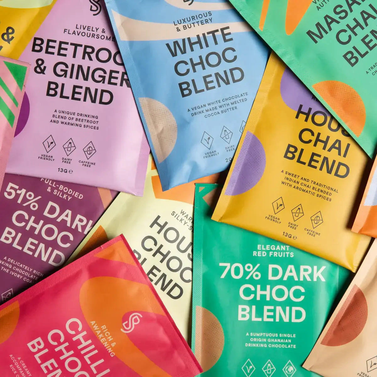 A vibrant display of Blendsmiths' Curiosity Collection, a selection of premium drinking chocolate, matcha chai, and superfood blends. A hand is holding a Japanese Matcha Blend sachet, while a stack of other flavored sachets, including Original Choc Blend,