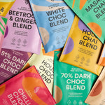 A vibrant display of Blendsmiths' Curiosity Collection, a selection of premium drinking chocolate, matcha chai, and superfood blends. A hand is holding a Japanese Matcha Blend sachet, while a stack of other flavored sachets, including Original Choc Blend,