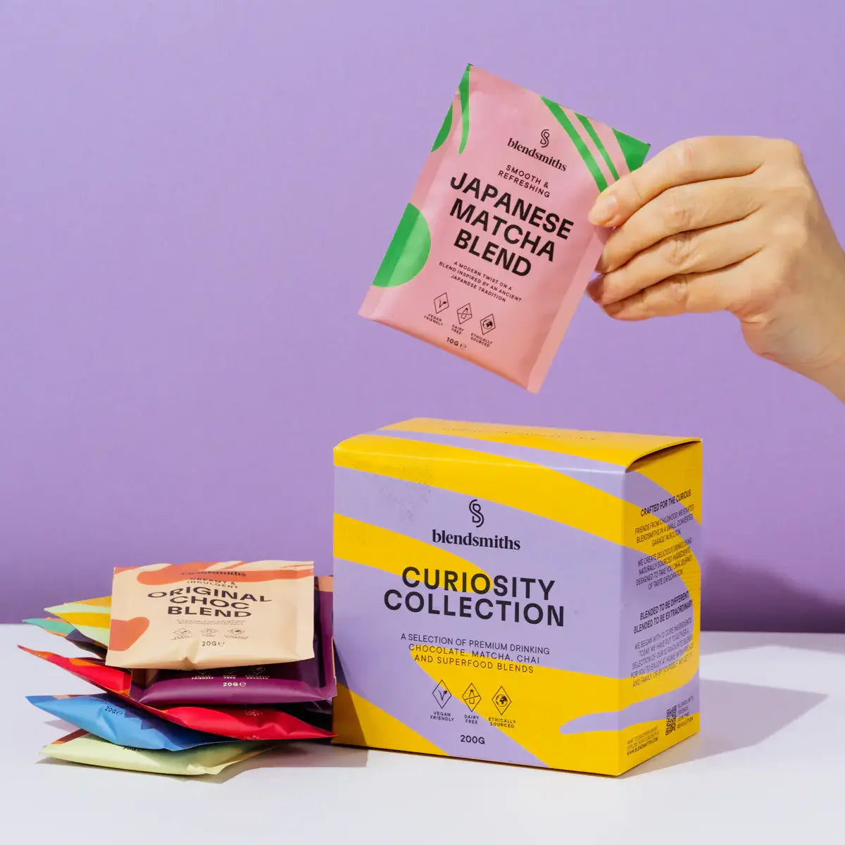 A vibrant display of Blendsmiths' Curiosity Collection, a selection of premium drinking chocolate, matcha chai, and superfood blends. A hand is holding a Japanese Matcha Blend sachet, while a stack of other flavored sachets, including Original Choc Blend,