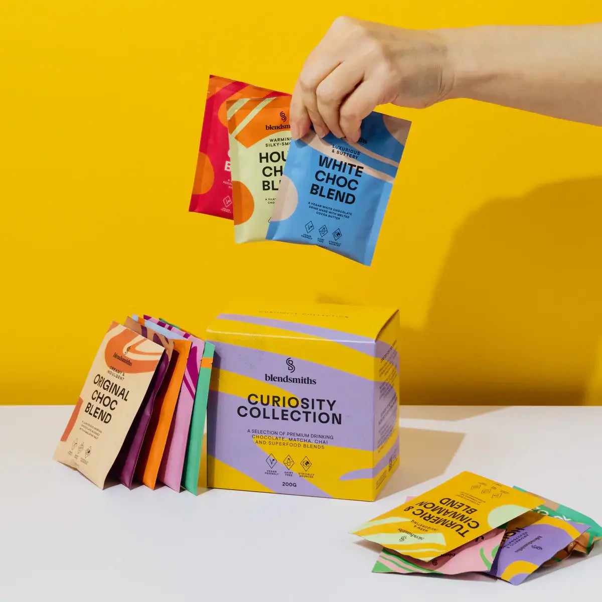 A vibrant display of Blendsmiths' Curiosity Collection, a selection of premium drinking chocolate, matcha chai, and superfood blends. A hand is holding a Japanese Matcha Blend sachet, while a stack of other flavored sachets, including Original Choc Blend,
