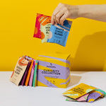 A vibrant display of Blendsmiths' Curiosity Collection, a selection of premium drinking chocolate, matcha chai, and superfood blends. A hand is holding a Japanese Matcha Blend sachet, while a stack of other flavored sachets, including Original Choc Blend,