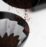 Pouring ground coffee into a FLO 55 Paper Filters 100x, ideal for rich and bright brew options.