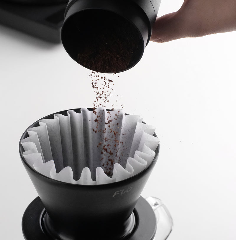 Pouring coffee grounds into a Varia FLO dripper with FLO 85 Paper Filters 100x for optimal brewing.