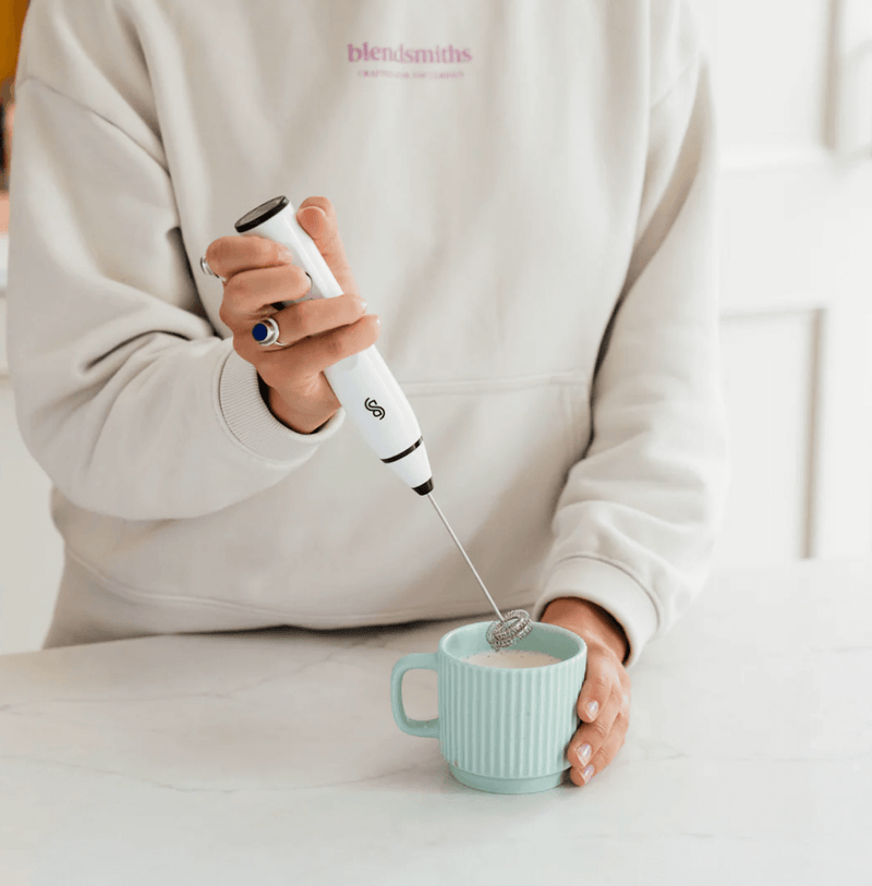 Person using the Blendsmiths Handheld Milk Frother to create foam in a ceramic cup in a kitchen setting.
