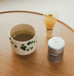 Hojicha tea setup with a ceramic cup, whisk, and tin on a wooden table, showcasing the warm, nutty flavor of roasted Japanese green tea.