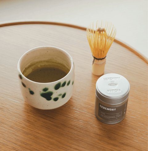 Hojicha tea setup with a ceramic cup, whisk, and tin on a wooden table, showcasing the warm, nutty flavor of roasted Japanese green tea.