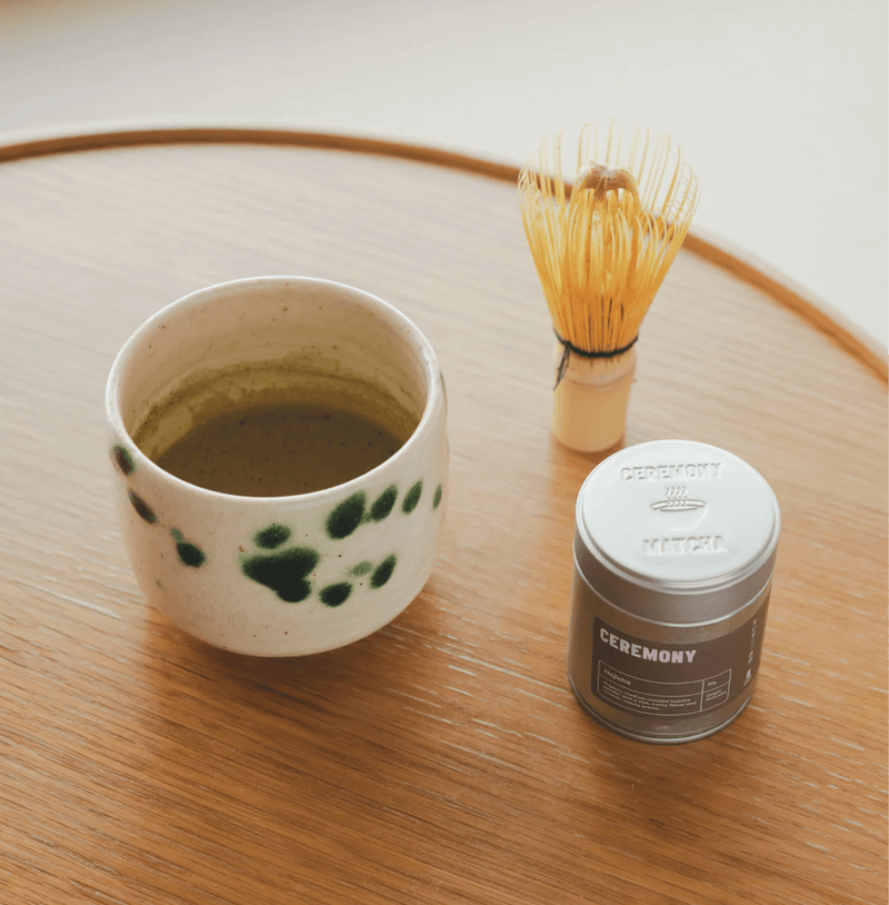 Hojicha tea setup with a ceramic cup, whisk, and tin on a wooden table, showcasing the warm, nutty flavor of roasted Japanese green tea.