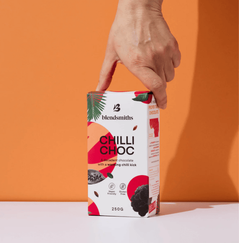 Hand holding a pack of Chilli Chocolate Blend with vibrant packaging against an orange background.