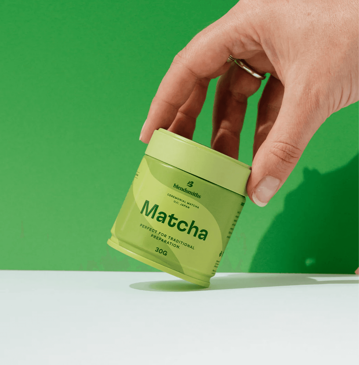 Hand holding a tin of Pure Ceremonial Grade Matcha against a green background.
