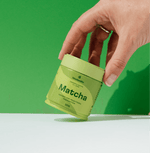 Hand holding a tin of Pure Ceremonial Grade Matcha against a green background.