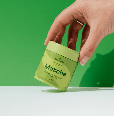Hand holding a tin of Pure Ceremonial Grade Matcha against a green background.
