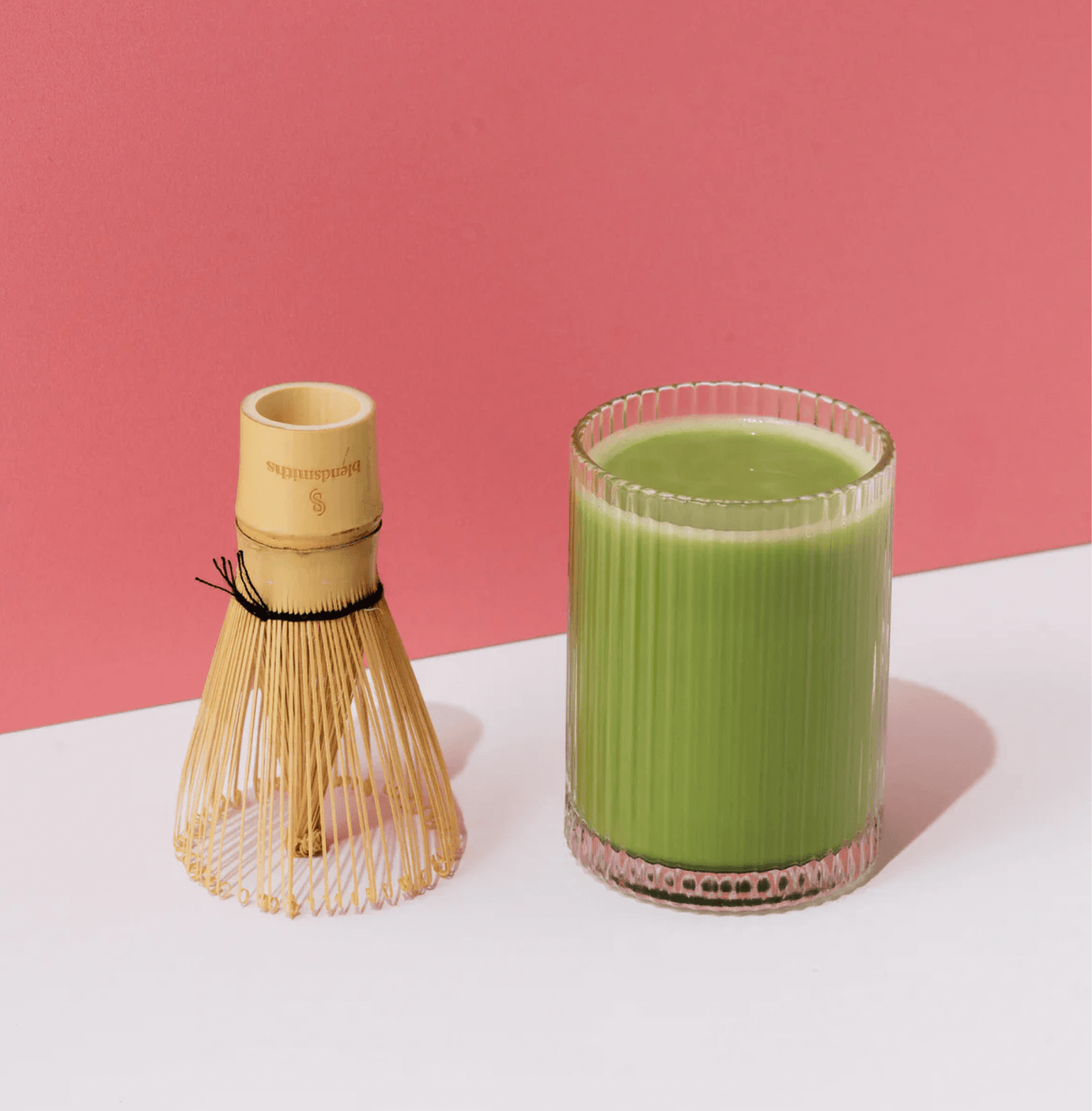 Glass of Wakaba Ceremonial Matcha with bamboo whisk against a pink background