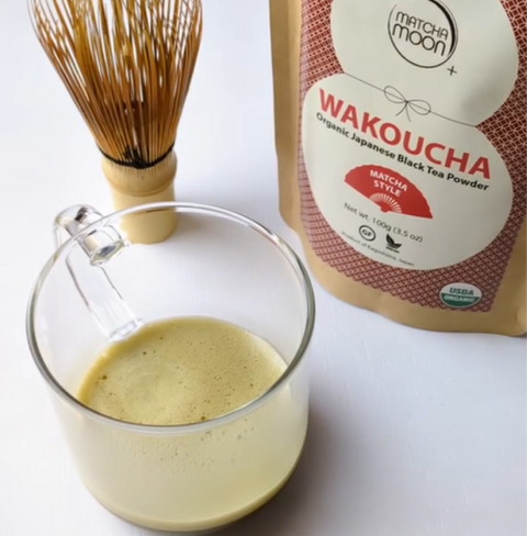 Cup of brewed Wakoucha Organic Hojicha Black Tea Powder next to a whisk and product packaging.