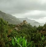 Scenic view of lush green hills and historic building enveloped in misty clouds, depicting vibrant natural landscape.