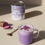 Purple UAE Fog latte with creamy ube blend in glass mug next to product can and sliced purple yam on beige surface.