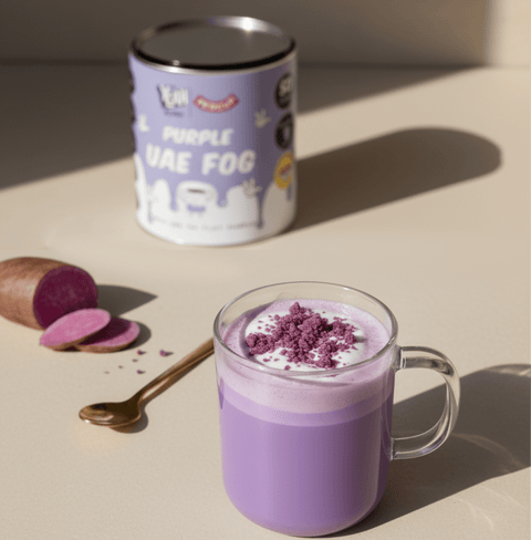 Purple UAE Fog latte with creamy ube blend in glass mug next to product can and sliced purple yam on beige surface.