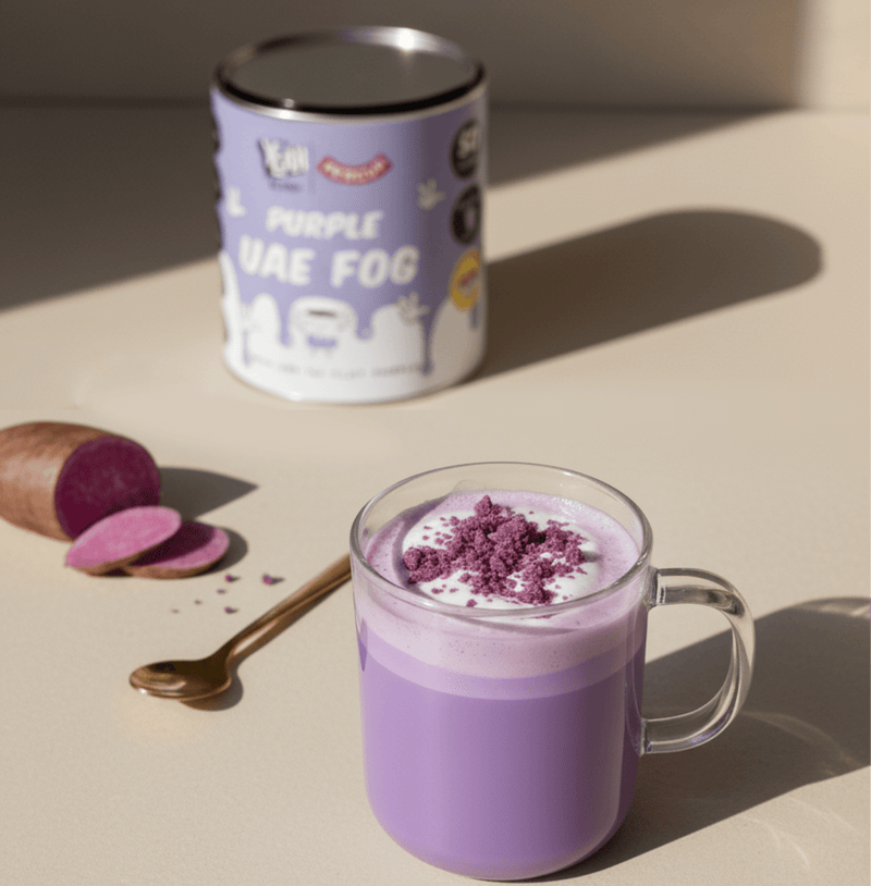 Purple UAE Fog latte with creamy ube blend in glass mug next to product can and sliced purple yam on beige surface.