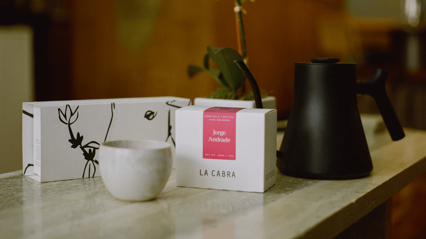 Ceramic cup, black kettle, and white box with branding on a wooden surface.