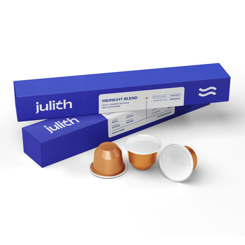 Julith Midnight Blend coffee capsules shown with their blue packaging boxes on a clean white background. The image features three coffee pods: one standing upright showing its metallic bronze cup, one lying down showing its white interior, and a third cap off, revealing the white coffee reservoir.