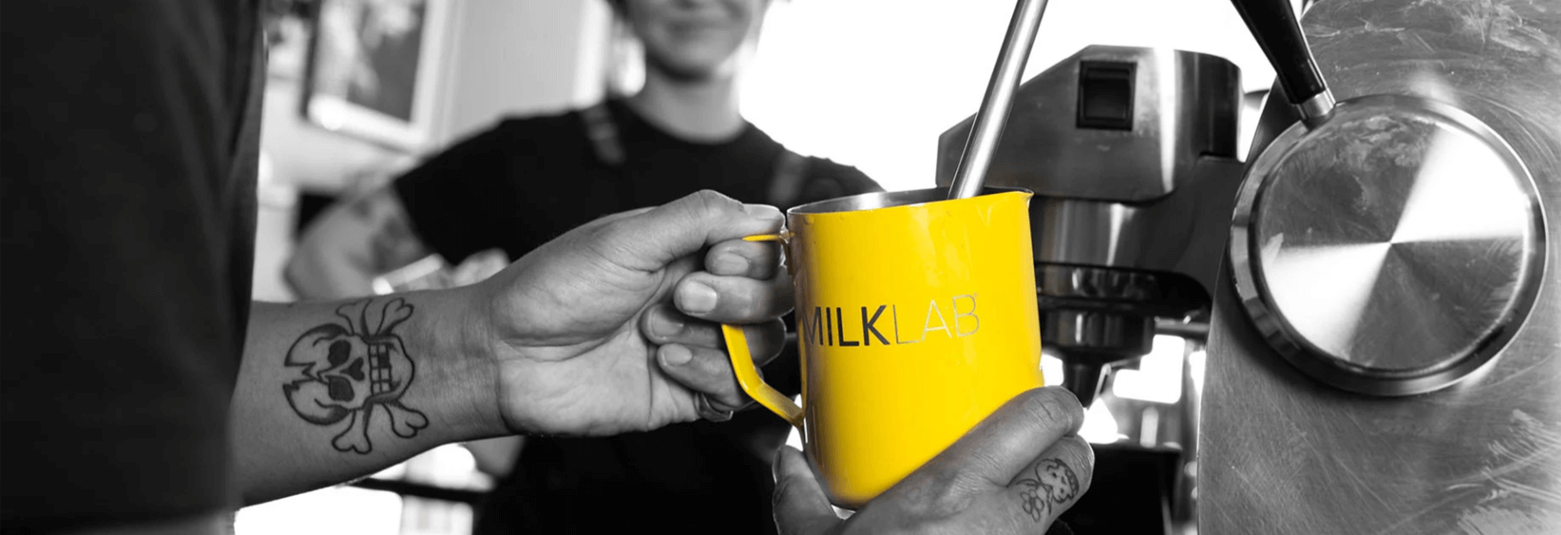 Barista pouring coffee into a bright yellow MILKLAB mug, showcasing drinkware accessories in a cafe.