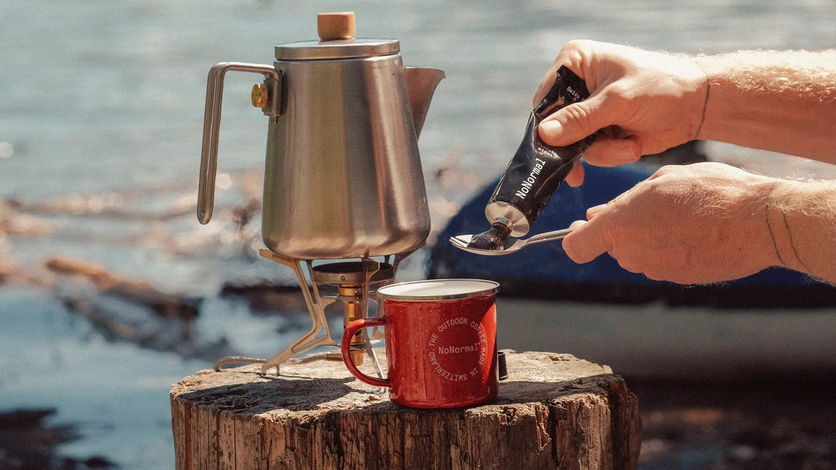 A person's hands are shown lighting a small camping stove with a black lighter, with a stainless steel coffee pot and a red enamel mug sitting on a rustic wooden log. The background features a scenic lake and mountains.