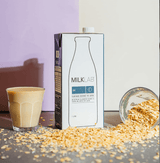 Oat Milk from MILKLAB in a carton next to a glass and oats on a table, creamy plant-based drink for coffee.