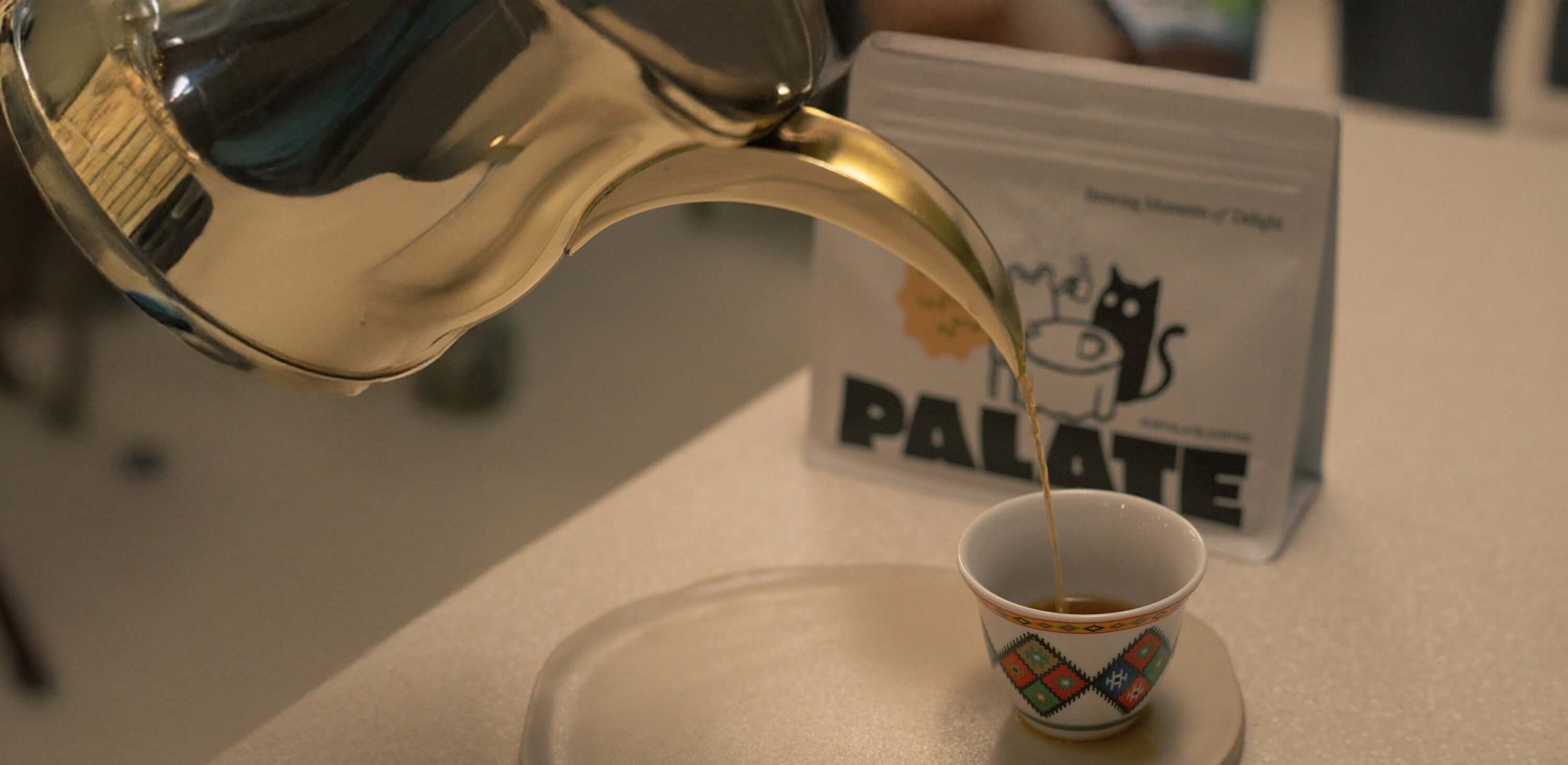 Golden kettle pouring fresh coffee into an elegant cup, with Palate coffee beans in the background.