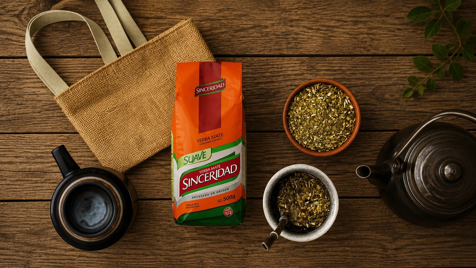 A bag of Sinceroíad Suave whole green tea leaves is displayed on a rustic wooden table alongside a jute tote bag, a black ceramic teapot, a white bowl with loose tea and a spoon, a small terracotta bowl of dried yerba mate, and green leaves.