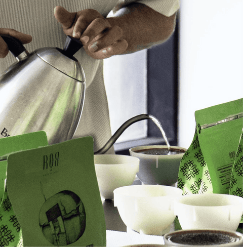 Person brewing coffee with green ROR coffee bags and cups on a table.