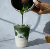 Green Matcha being poured into a clear cup with a white lid on a light gray background