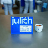 A blueJulith branded card or box stands on a gray tabletop next to a small white cup filled with a reddish-brown beverage. The background is softly blurred with people visible, indicating a real environment.