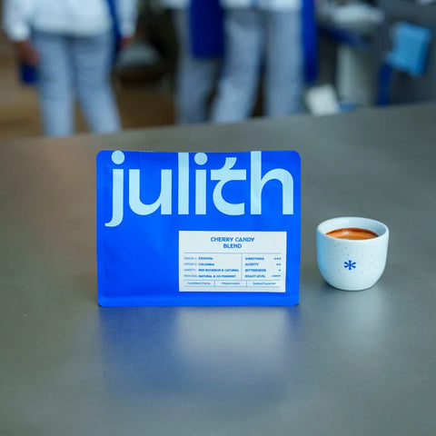 A blueJulith branded card or box stands on a gray tabletop next to a small white cup filled with a reddish-brown beverage. The background is softly blurred with people visible, indicating a real environment.
