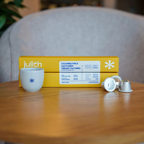 Julith Colombian fine coffee capsules, featuring a yellow box labeled 'COLOMBIA FINCA LAS FLORES CREAMY CULTURED', a small white ceramic cup with a blue flower, and two silver coffee capsules on a wooden table in front of a blurred gray sofa.
