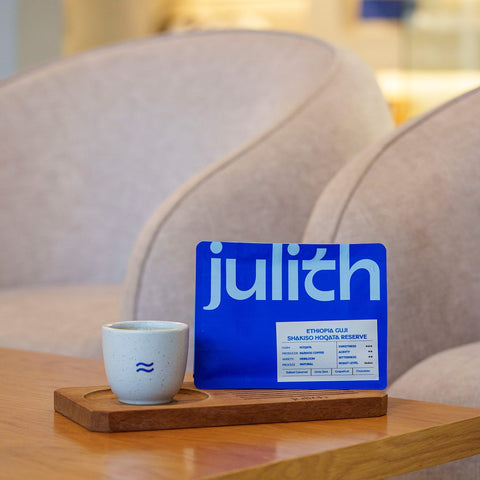 Blue coffee bag with 'julith' branding on a wooden coaster next to a white cup on a wooden table.