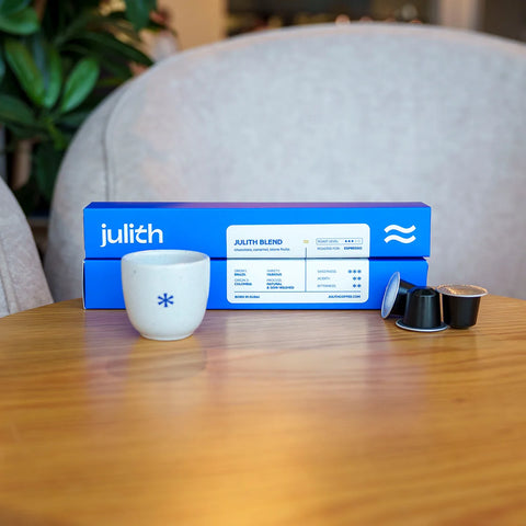 Julith coffee blend packaging with a blue box, a white ceramic cup with a blue star, and two coffee pods on a wooden table in a cozy indoor setting.