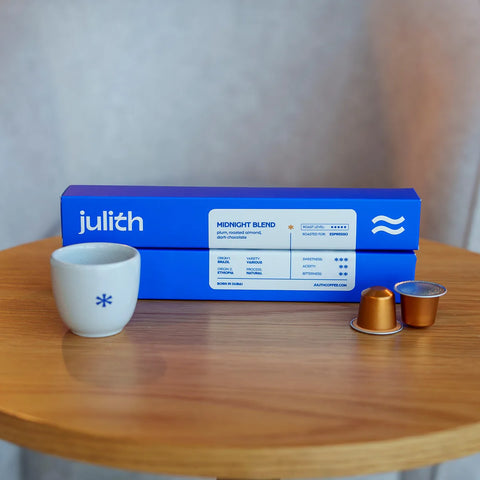 Julith Midnight Blend coffee capsules shown with their blue retail box on a wooden table, alongside a small white ceramic cup and two individual coffee pods.