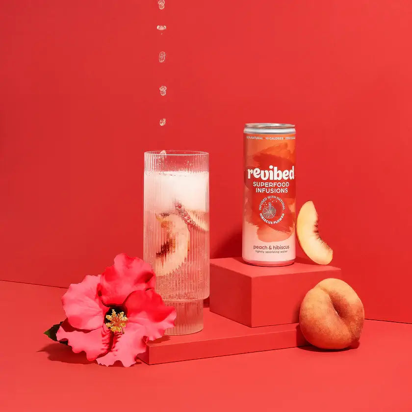 Can of Hibiscus & Peach Superfood Infusion with fresh peach slices and hibiscus flower on a red backdrop.