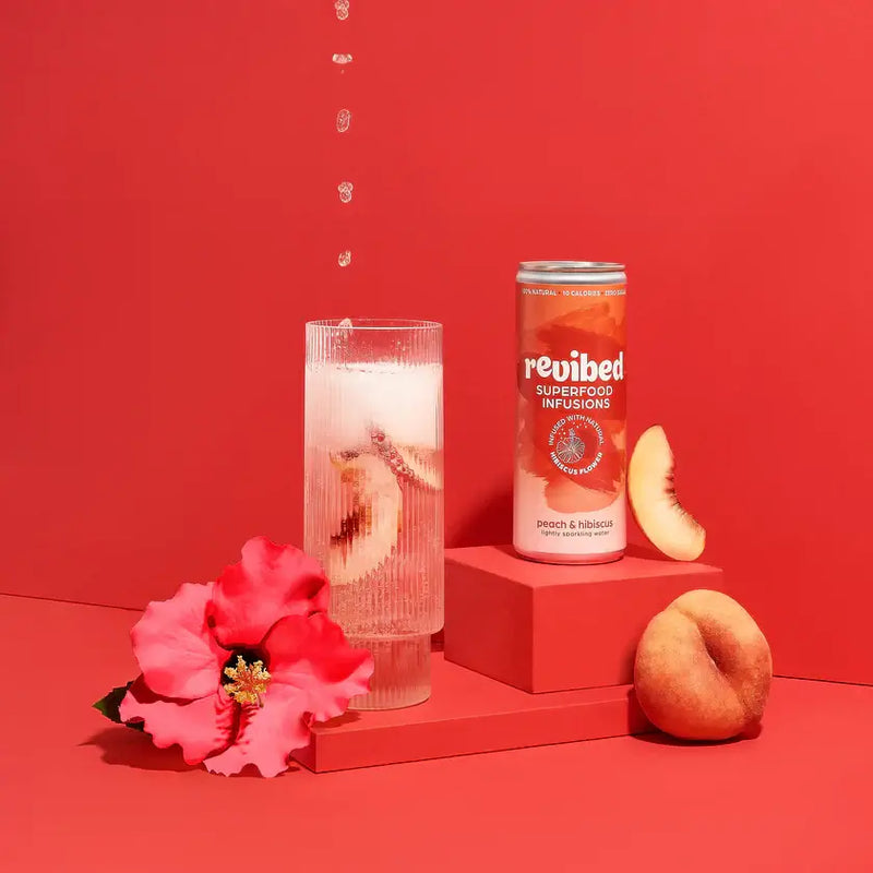 Can of Hibiscus & Peach Superfood Infusion with fresh peach slices and hibiscus flower on a red backdrop.