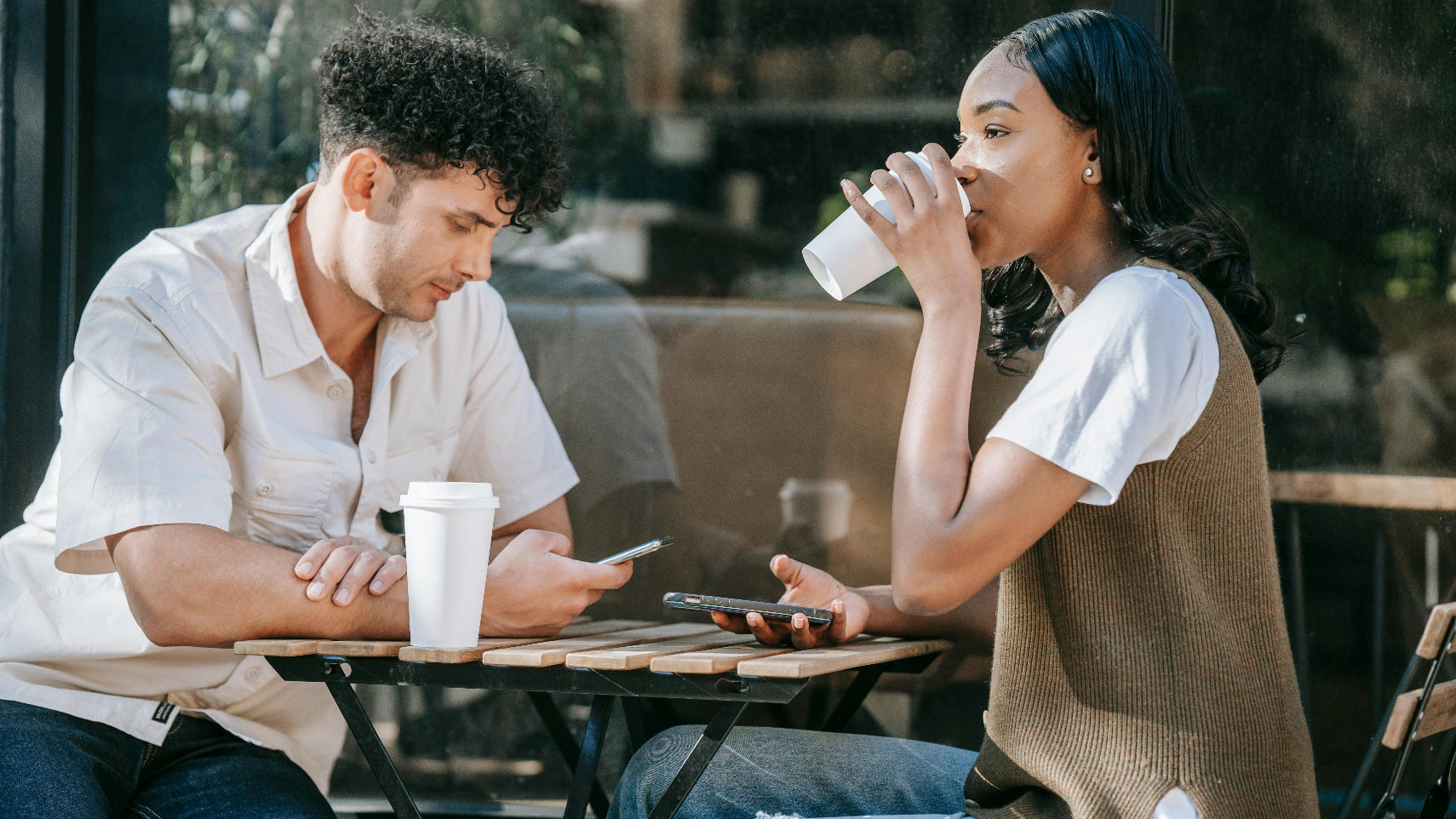 Two people sitting at a table outdoors, one using a phone and the other drinking from a cup.
