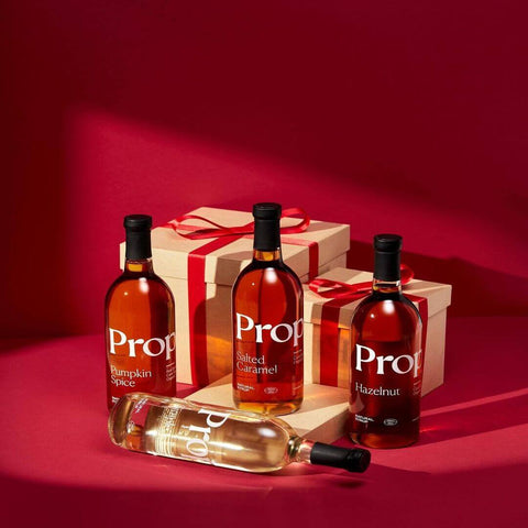 Three bottles of Proper Natural Syrups, including Salted Caramel Syrup, on a festive red background.