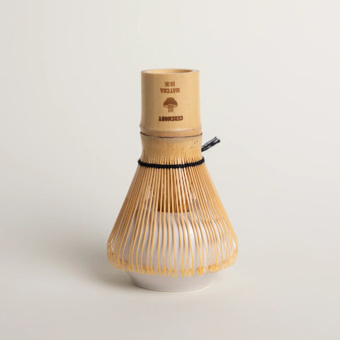 A Japanese matcha whisk (chashaku) made of light natural bamboo and tan string, featuring a white ceramic base and a black rubber stopper, set against a clean off-white background.