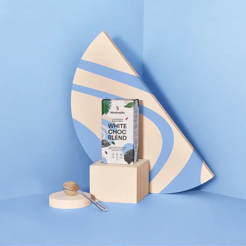 Blendsmiths White Choc Blend packaging, featuring a luxurious and buttery vegan white chocolate drink made with melted cocoa butter. The box has a sleek design with soft blue and earthy tones, abstract patterns, and botanical elements. 