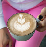 Cup of organic Hojicha with latte art held by a person in pink pants, showcasing Japanese roasted green tea.